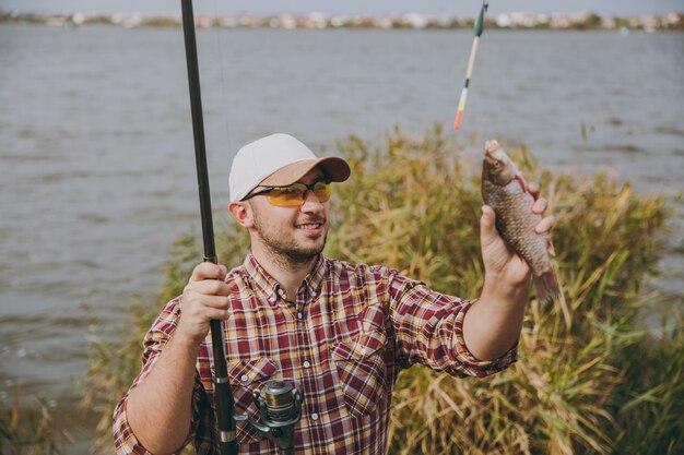 Photo young unshaven smiling man in checkered shirt, cap and sunglasses pulled out fishing pole and holds caught fish on shore of lake near shrubs and reeds. lifestyle, recreation, fisherman leisure concept