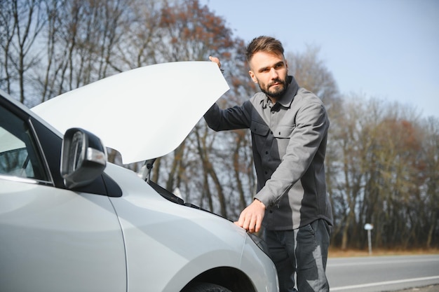 Young upset casual man trying to fix his broken car outdoors Man waiting for towing service for help car accident on the road Roadside assistance concept