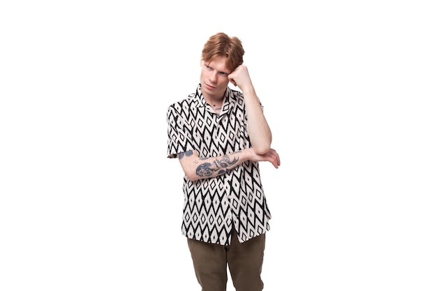 Young upset caucasian man with red hair and a tattoo is dressed in a shirt with a pattern of rhombus