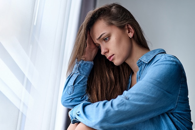 Photo young upset depressed lonely pensive woman with sad eyes in a shirt holds his head with hand sits alone at home by the window during the depression and worries