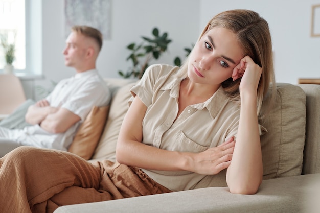 Young upset and lonely wife sitting on couch with her hand by head while expressing offense and misunderstanding during quarrel with husband