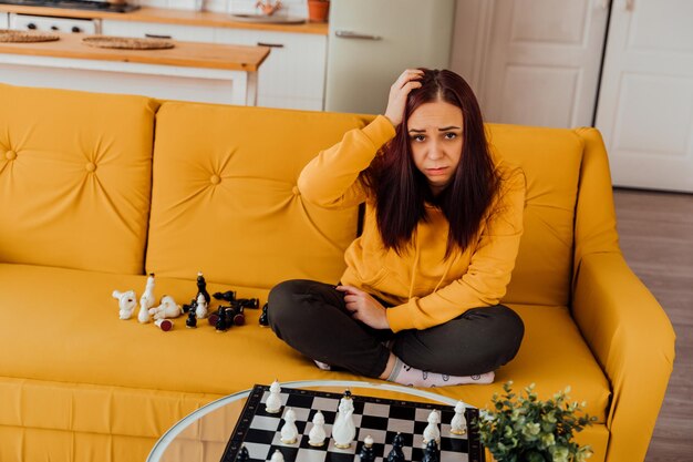 Young upset woman playing chess sitting on sofa Distressed female plays in logical board game with herself in room