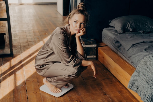 Young upset woman in satin pajama measuring her weight at home