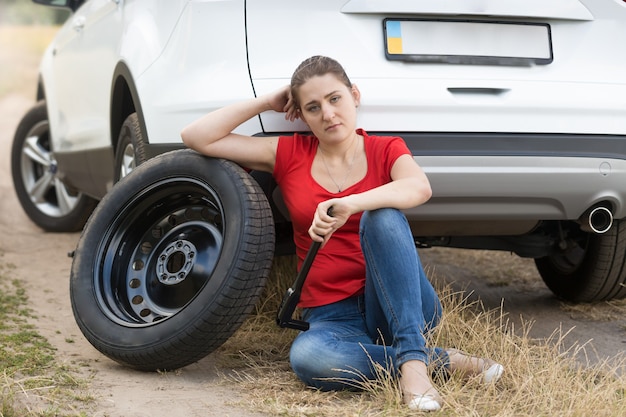 壊れた車に寄りかかって地面に座っている若い動揺女性