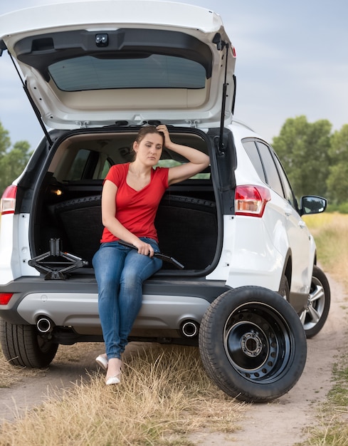 開いた車のトランクに座って、タイヤを交換する助けを待っている若い動揺の女性