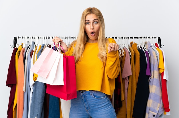 Young Uruguayan blonde woman in a clothing store and holding shopping bags surprised and pointing front