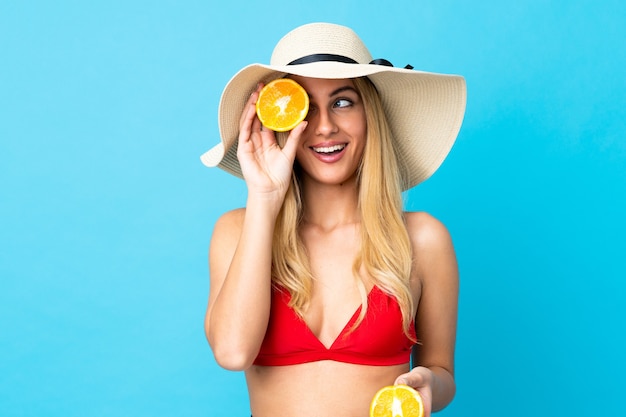 Young Uruguayan blonde woman over isolated blue background in swimsuit and holding an orange