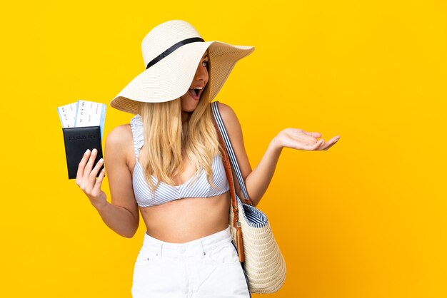 Young Uruguayan blonde woman in swimsuit holding a passport over isolated yellow background with surprise facial expression