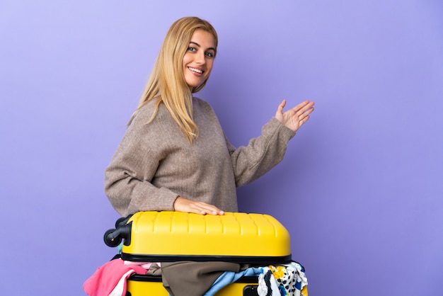 Молодая уругвайская белокурая женщина с чемоданом, полным одежды над изолированной фиолетовой стеной, протягивающей руки в сторону для приглашения прийти