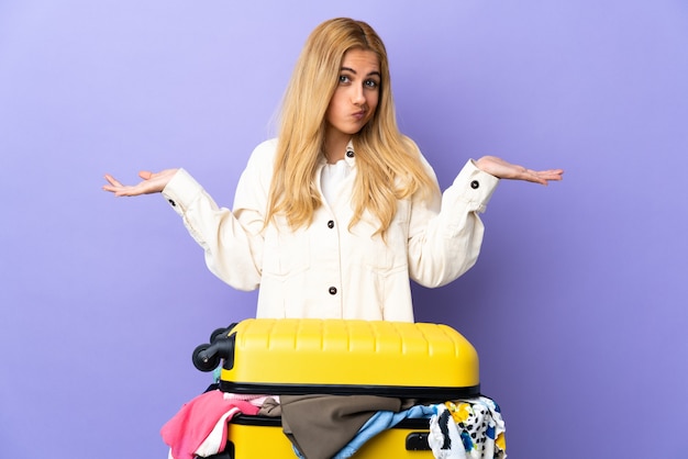 Young Uruguayan blonde woman with a suitcase full of clothes over isolated purple wall having doubts while raising hands