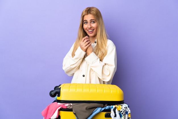 Young Uruguayan blonde woman with a suitcase full of clothes over isolated purple wall laughing