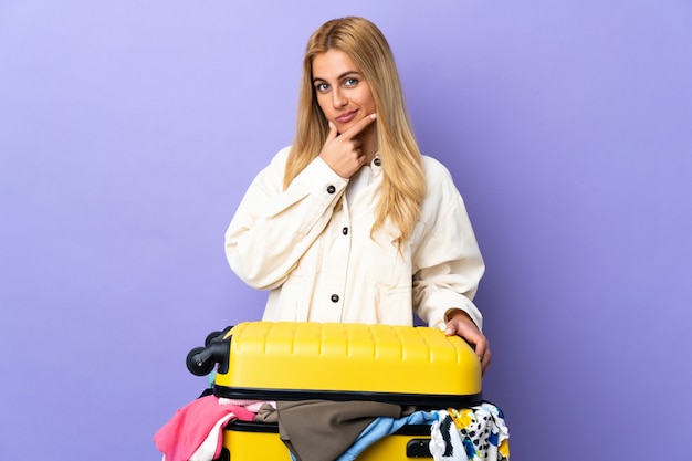 Young Uruguayan blonde woman with a suitcase full of clothes over isolated purple wall thinking