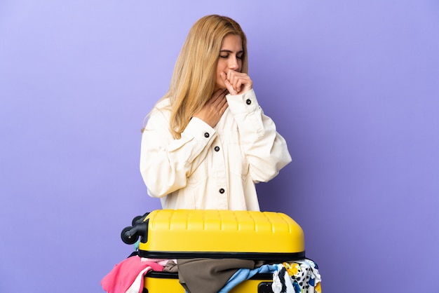 Young Uruguayan blonde woman with a suitcase full of clothes over purple wall coughing a lot