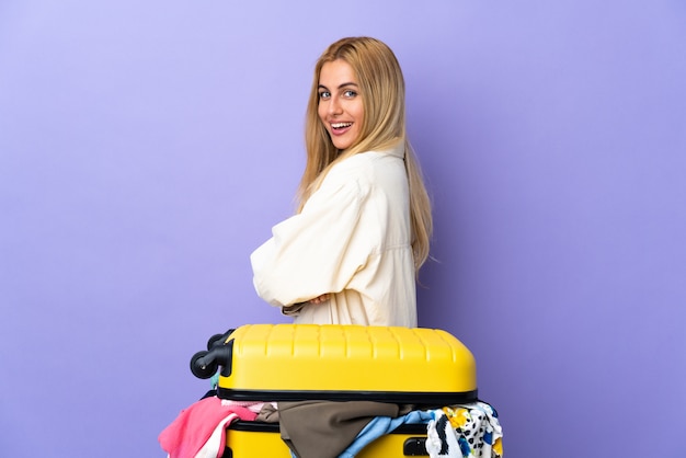 Photo young uruguayan blonde woman with a suitcase full of clothes over purple wall in lateral position