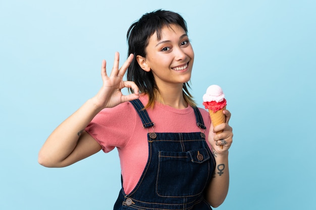 Young Uruguayan girl holding a cornet ice cream over isolated blue wall showing ok sign with fingers