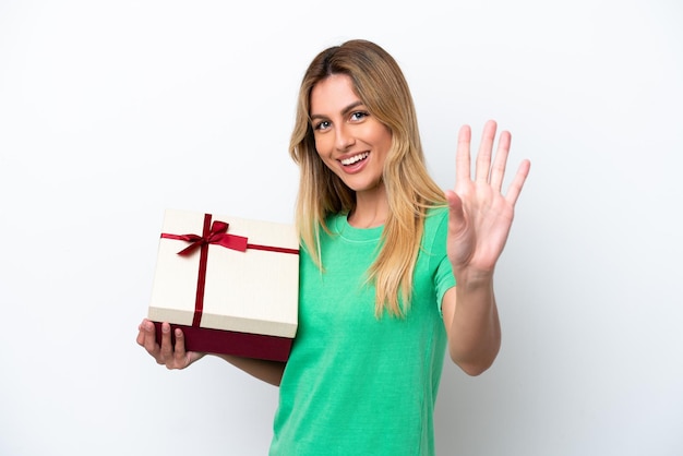 Young Uruguayan woman holding a gift isolated on white background saluting with hand with happy expression