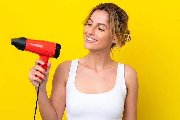 Young Uruguayan woman holding a hairdryer isolate don yellow background with happy expression