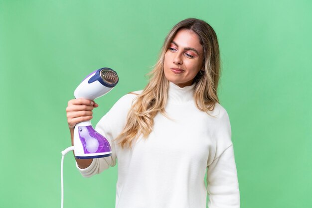 Young Uruguayan woman holding an iron over isolated background with sad expression