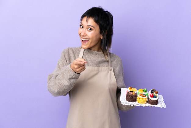 Young Uruguayan woman holding lots of different mini cakes on isolated purple points finger at you