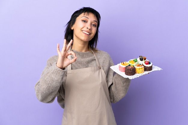 손가락으로 확인 표시를 보여주는 고립 된 보라색에 다른 미니 케이크를 많이 들고 젊은 우루과이 여자