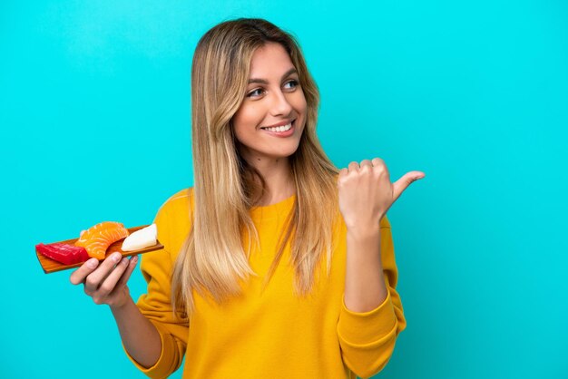 Young Uruguayan woman holding sashimi isolated on blue background pointing to the side to present a product