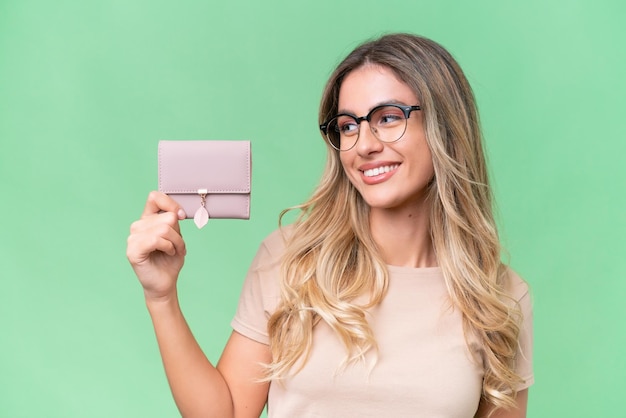 Young Uruguayan woman holding a wallet over isolated background with happy expression