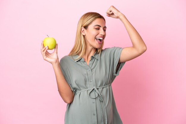 Young Uruguayan woman isolated on blue background pregnant and holding an apple while celebrate a victory