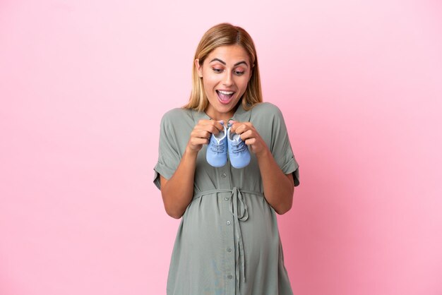 Young Uruguayan woman isolated on blue background pregnant and holding baby booties with surprised expression