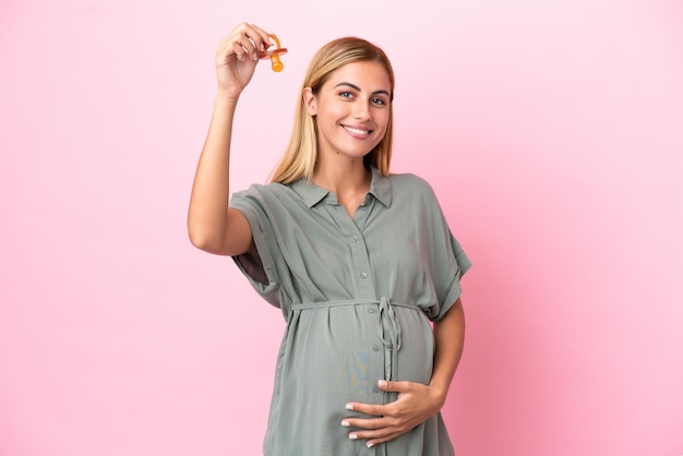 Young Uruguayan woman isolated on blue background pregnant and holding a pacifier