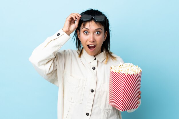 Young Uruguayan woman over isolated blue surprised with 3d glasses and holding a big bucket of popcorns