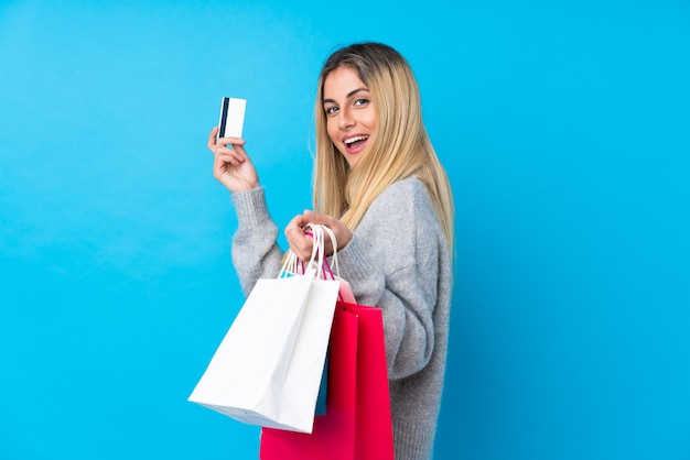 Young Uruguayan woman over isolated blue wall holding shopping bags and a credit card