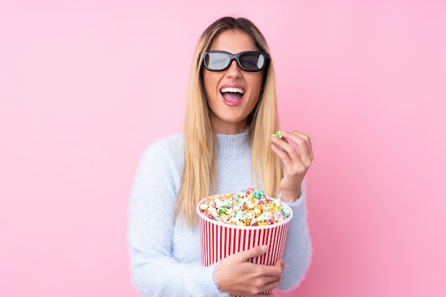 Young Uruguayan woman over isolated pink wall with 3d glasses and holding a big bucket of popcorns while pointing front