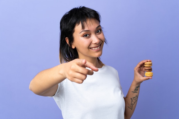 Молодая уругвайская женщина над изолированной фиолетовой стеной держит красочные французские макаруны и указывает пальцем на тебя