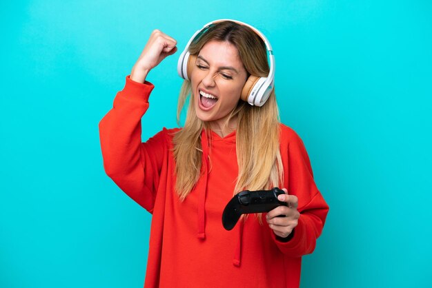 Young Uruguayan woman playing with a video game controller isolated on blue celebrating a victory