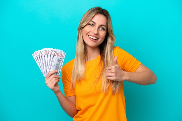 Young Uruguayan woman taking a lot of money isolated on blue background giving a thumbs up gesture