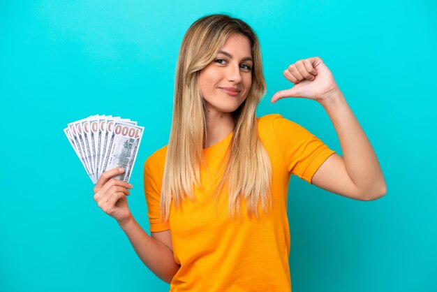 Young uruguayan woman taking a lot of money isolated on blue background proud and selfsatisfied