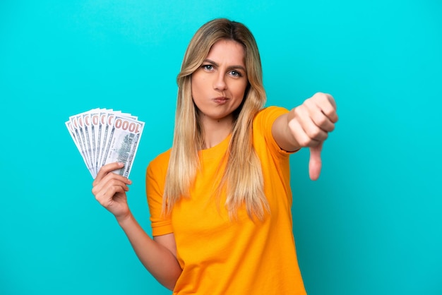 Young Uruguayan woman taking a lot of money isolated on blue background showing thumb down with negative expression