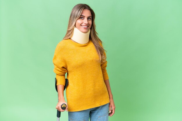 Young Uruguayan woman wearing neck brace and crutches over isolated background smiling a lot