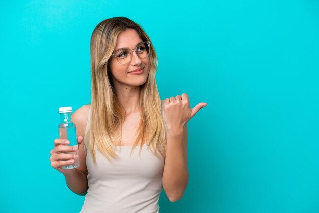 Young Uruguayan woman with a bottle of water isolated on blue background pointing to the side to present a product