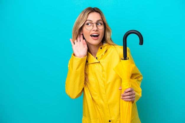 Young Uruguayan woman with rainproof coat and umbrella isolated on blue background listening to something by putting hand on the ear