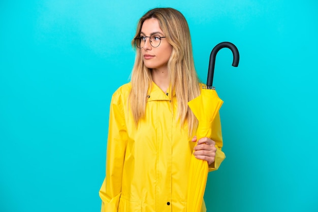 Young Uruguayan woman with rainproof coat and umbrella isolated on blue background looking to the side