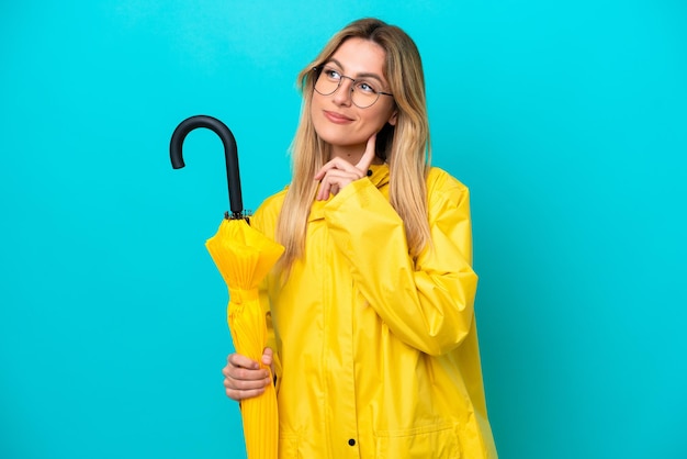 Young Uruguayan woman with rainproof coat and umbrella isolated on blue background thinking an idea while looking up