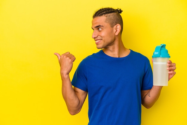 Young venezuelan man drinking a protein shake isolated on yellow background points with thumb finger away, laughing and carefree.