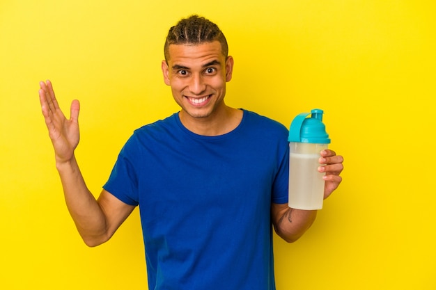 Young venezuelan man drinking a protein shake isolated on yellow background receiving a pleasant surprise, excited and raising hands.