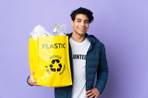 Young Venezuelan man holding a bag full of plastic bottles posing with arms at hip and smiling