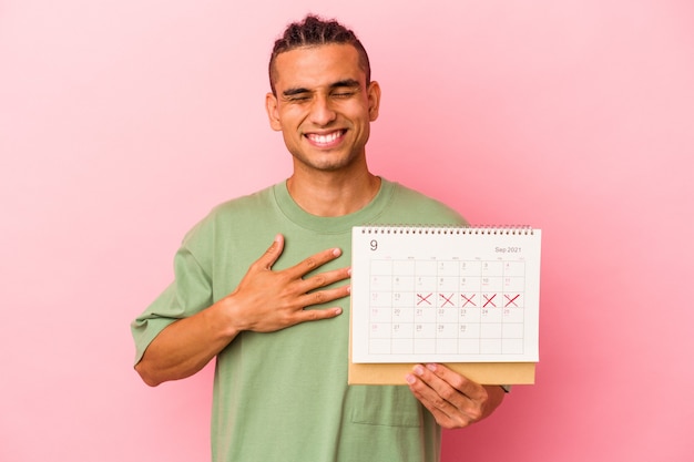 Young venezuelan man holding a calendar isolated on pink wall laughs out loudly keeping hand on chest.