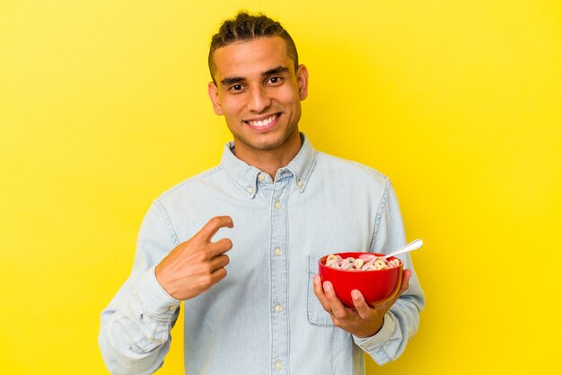 Young venezuelan man holding a cereals bowl isolated on yellow background pointing with finger