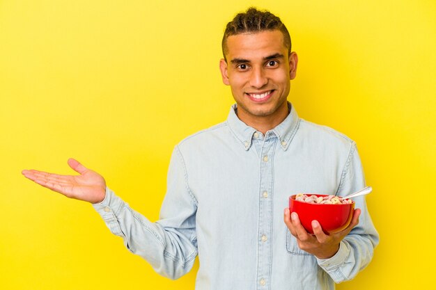 Young venezuelan man holding a cereals bowl isolated on yellow background showing a copy space on a palm and holding another hand on waist.