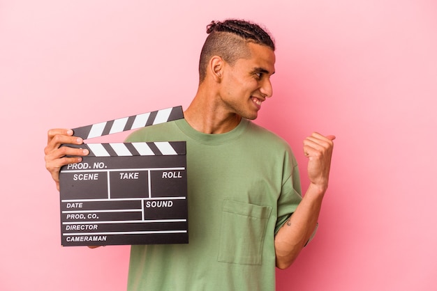 Young venezuelan man holding a clapperboard isolated on pink background points with thumb finger away, laughing and carefree.