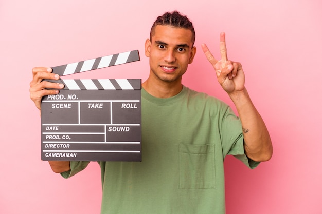 Young venezuelan man holding a clapperboard isolated on pink background showing number two with fingers.